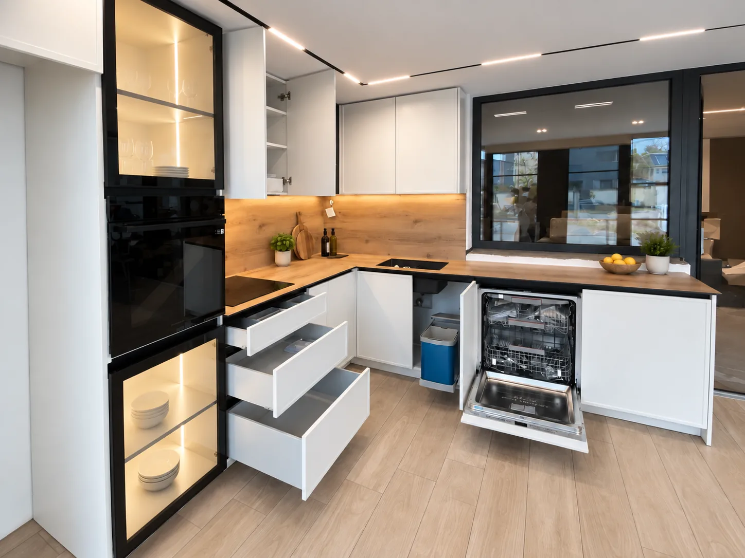 Handleless white kitchen with oak worktop, black-framed glazed cabinetry and lit interiors — drawer and appliance storage
