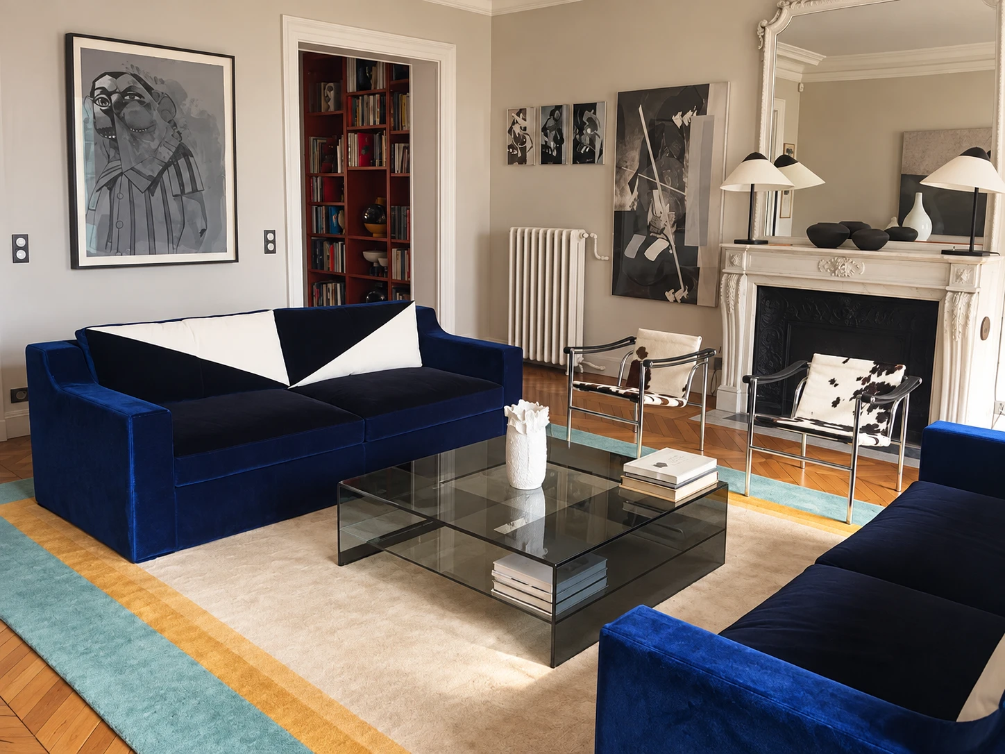 A sitting room with a marble fireplace: two deep blue velvet sofas face each other across a glass coffee table on a striped rug